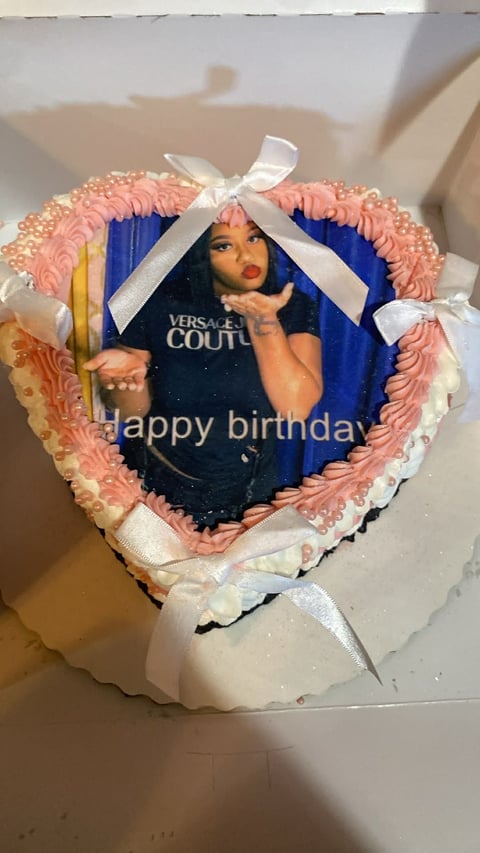 Heart-shaped birthday cake with pink frosting, white ribbon bow, and printed photo of person wearing Versace Couture shirt